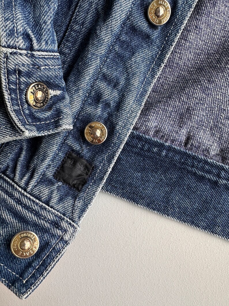 May include: Close-up of a blue denim jacket with gold metal buttons. The buttons have the text "Versace Jeans Couture" engraved on them.