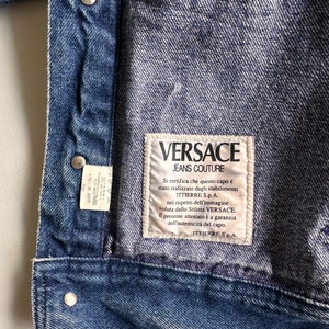May include: A blue denim jacket with a white label that reads "VERSACE JEANS COUTURE". The label has text in Italian that certifies the authenticity of the garment.