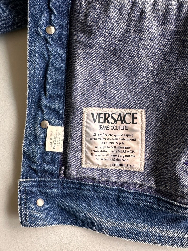 May include: A blue denim jacket with a white label that reads "VERSACE JEANS COUTURE". The label has text in Italian that certifies the authenticity of the garment.