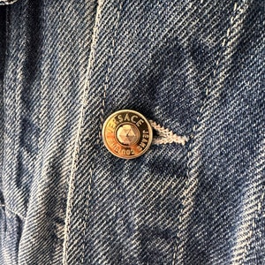 May include: Close-up of a blue denim jacket with a gold metal button that reads "VERSACE JEANS COUTURE".