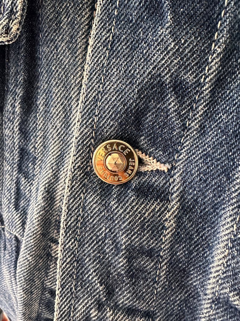 May include: Close-up of a blue denim jacket with a gold metal button that reads "VERSACE JEANS COUTURE".