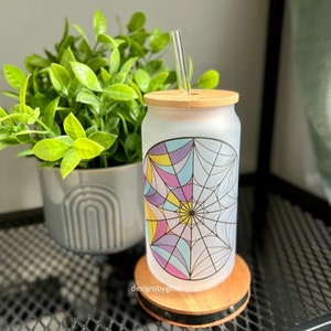 May include: A frosted glass tumbler with a colorful geometric spiderweb design and a bamboo lid. The tumbler has a straw and is sitting on a wooden coaster.
