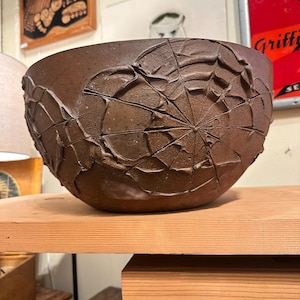 May include: A brown ceramic bowl with a textured spiderweb design. The bowl is round and sits on a wooden shelf. The spiderweb design is raised and adds a unique, artistic element to the piece. This decorative bowl is perfect for home decor.