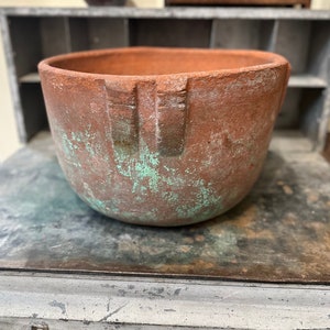 Könnte beinhalten: Ein Terrakotta-Pflanztopf mit einer abgenutzten, gealterten Oberfläche. Der Topf hat eine breite, runde Öffnung und eine leicht ausgestellte Lippe. Die Oberfläche ist bräunlich mit grünen Akzenten.