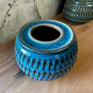 Op de afbeelding: Een tealblauwe keramische plantenpot met een getextureerd, verhoogd patroon. De pot heeft een brede, ronde opening en een licht afgeronde basis.