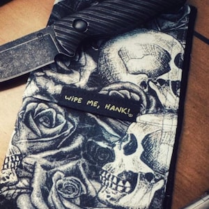 May include: A black and white patterned knife sheath with a skull and rose design. The sheath has the text "WIPE ME, HANK!" and a black knife with a textured handle.