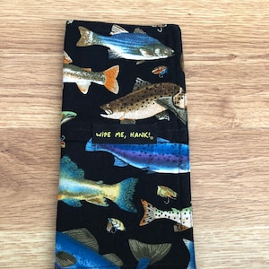 Può includere: Tessuto nero con un motivo ripetuto di pesci colorati e esche da pesca. Il tessuto ha un'etichetta che dice "Wipe Me, Hank!"