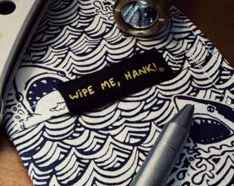 May include: A pocket knife, a brass flashlight, and a pen rest on a navy and white patterned cloth with shark illustrations. The cloth has the text "WIPE ME, HANK!" in yellow.