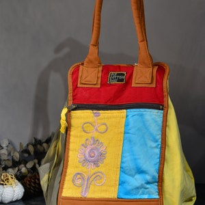 May include: A multi-colored patchwork tote bag with brown leather handles. The bag has a yellow, blue, and red panel with embroidered flowers. The bag has a brown leather patch with the text "OLD COTTON BAGS".