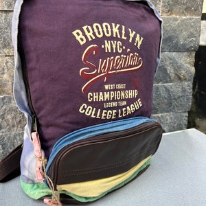 Może przedstawiać: Fioletowy plecak z płótna z brązową klapą skórzaną i kolorowym paskowanym wzorem. Plecak ma nadrukowany tekst "Brooklyn NYC Superior West Coast Championship Legend Team College League" na przodzie.