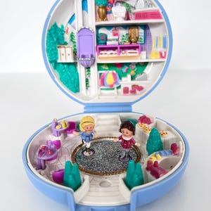 Könnte beinhalten: Ein geöffnetes, blau-weißes Polly Pocket Spielset, das eine Miniatur-Salon-Szene zeigt. Es enthält zwei kleine Figuren, eine Saloneinrichtung mit Regalen und dekorativen Elementen. Das Spielset ist ein nostalgisches Spielzeug.