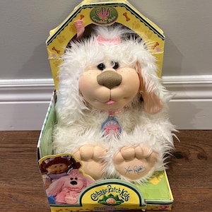 May include: A Cabbage Patch Kids Lovable Pets plush dog toy in its original box. The white plush dog has a pink bow, a brown nose, and a pink tongue. The box is yellow and green with the Cabbage Patch Kids logo.