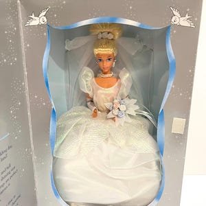 May include: A white bridal doll with blonde hair, wearing a white gown with a blue ribbon and holding a bouquet of blue and white flowers. The doll is in a clear plastic box with a blue ribbon and a silver background.