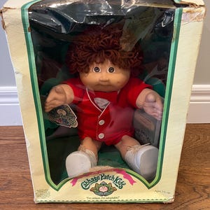 Peut inclure: Une poupée Cabbage Patch Kids dans sa boîte d'origine. La poupée a des cheveux bruns bouclés, une tenue rouge et des chaussures blanches. La boîte est de couleur crème avec des accents verts et le logo Cabbage Patch Kids. Le texte sur la boîte indique "I'm unique, I'm adoptable!" et "Ages 3 & up."
