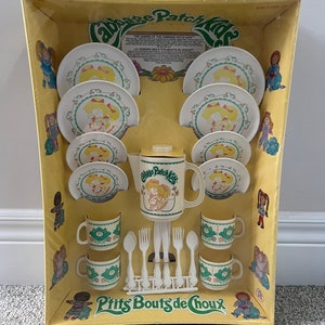 Peut inclure: Un coffret de thé Cabbage Patch Kids dans son emballage d'origine. L'ensemble comprend une théière, des tasses, des soucoupes et une fourchette et une cuillère. L'emballage est jaune avec des accents verts et blancs. Le texte sur l'emballage indique "Cabbage Patch Kids" et "P'tits Bouts de Choux".
