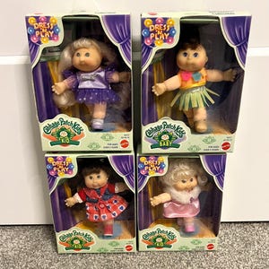Peut inclure: Quatre poupées Cabbage Patch Kids dans leurs boîtes d'origine. Chaque poupée porte une tenue différente, dont une robe violette, une jupe verte, une robe rouge et une robe rose. Les boîtes présentent le logo "Dress 'N Play" et sont étiquetées "Kid" ou "Baby".