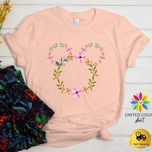 May include: A pink t-shirt with a floral design in the shape of Mickey Mouse ears. The design features pink, orange, and green flowers.