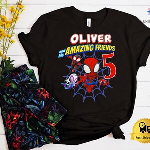 Pode incluir: Uma t-shirt preta de manga curta com uma impress&atilde;o gr&aacute;fica de tr&ecirc;s super-her&oacute;is de desenhos animados numa teia de aranha azul. O texto diz OLIVER and his AMAZING FRIENDS com um grande n&uacute;mero 5 vermelho. O design inclui cores vermelho, azul e branco.