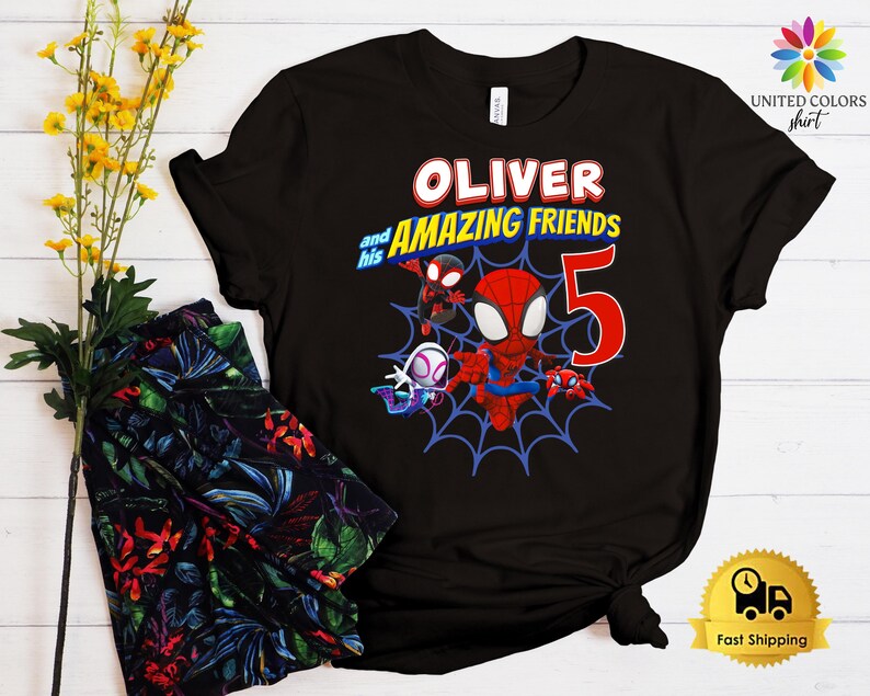 Pode incluir: Uma t-shirt preta de manga curta com uma impress&atilde;o gr&aacute;fica de tr&ecirc;s super-her&oacute;is de desenhos animados numa teia de aranha azul. O texto diz OLIVER and his AMAZING FRIENDS com um grande n&uacute;mero 5 vermelho. O design inclui cores vermelho, azul e branco.