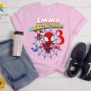 Pode incluir: Uma t-shirt rosa claro com um gr&aacute;fico do Spiderman e amigos, o texto EMMA and her AMAZING FRIENDS e o n&uacute;mero 3. A t-shirt &eacute; apresentada ao lado de cal&ccedil;as de ganga azuis, um copo com padr&atilde;o e um par de sapatilhas pretas e brancas.