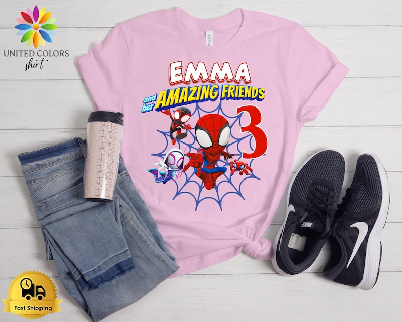 Pode incluir: Uma t-shirt rosa claro com um gr&aacute;fico do Spiderman e amigos, o texto EMMA and her AMAZING FRIENDS e o n&uacute;mero 3. A t-shirt &eacute; apresentada ao lado de cal&ccedil;as de ganga azuis, um copo com padr&atilde;o e um par de sapatilhas pretas e brancas.