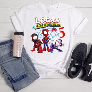 以下が含まれることがあります： スパイダーマンのキャラクターと「LOGAN and his AMAZING FRIENDS 5」の文字が描かれた、カラフルなグラフィックの白いTシャツ。デザインには、スパイダーマン、マイルズ・モラレス、スパイダーグウェンが含まれています。シャツは、青いジーンズ、トラベルマグ、黒いスニーカーと組み合わせています。