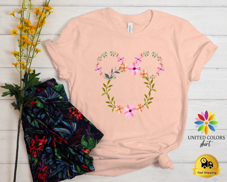 May include: A pink t-shirt with a floral design in the shape of Mickey Mouse ears. The design features pink, orange, and green flowers.