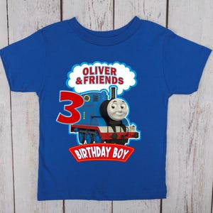 May include: A royal blue t-shirt featuring a graphic of a blue train with a smiling face. The shirt has the text "OLIVER & FRIENDS", the number "3", and the words "BIRTHDAY BOY" in a red banner. The shirt is designed for a child.