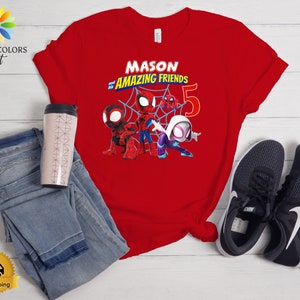 以下が含まれることがあります： スパイダーマンのキャラクターが描かれた赤いTシャツで、「MASON and AMAZING FRIENDS 5」の文字が入っています。画像には、青いジーンズ、トラベルマグ、黒いスニーカーが含まれています。子供服です。