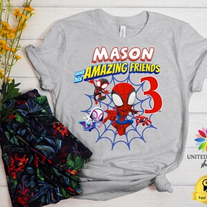 Pode incluir: Uma t-shirt cinzenta clara de manga curta com uma impress&atilde;o gr&aacute;fica com o texto MASON and his AMAZING FRIENDS e um grande n&uacute;mero 3 vermelho. O design inclui personagens de desenhos animados em vermelho, azul e branco sobre uma teia.