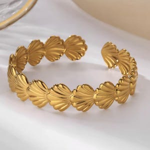 Peut inclure: Un bracelet manchette doré avec un motif de coquillage. Le bracelet est ouvert en haut et présente une série de formes de coquillages stylisées. Le bracelet est en métal brillant et réfléchissant.