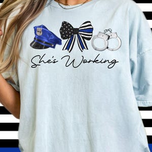 May include: Light blue t-shirt with a blue police hat, a blue and black ribbon, and silver handcuffs. The text "She's Working" is printed on the shirt.