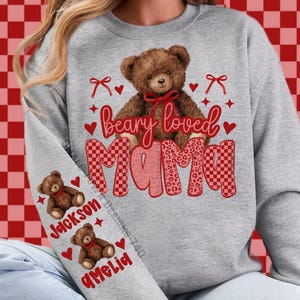 May include: A gray sweatshirt with a brown teddy bear graphic and the words "beary loved mama" in red and pink. The sleeve features two teddy bear graphics with the names "Jackson" and "Amelia". Hearts and bows are also included in the design.