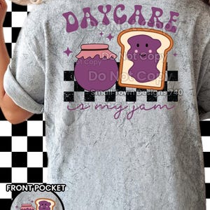 Puede incluir: Camiseta gris claro con un diseño lúdico. Presenta las palabras "DAYCARE is my jam" en escritura morada, un tarro de mermelada y una rebanada de pan tostado con mermelada, sobre un fondo a cuadros blanco y negro. Una versión más pequeña del diseño está en el bolsillo delantero.