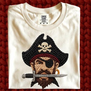 May include: Cream-colored t-shirt featuring a detailed pirate design. The pirate wears a black hat with a skull and crossbones, an eye patch, and a gold earring. A sword is held in the pirate's mouth. The shirt has a Comfort Colors tag.