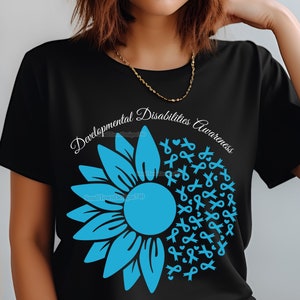 Puede incluir: Camiseta negra con un diseño de girasol azul y el texto "Developmental Disabilities Awareness". El girasol está hecho de formas de cinta azul.