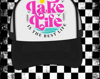 Könnte beinhalten: Weiße Trucker-Kappe mit schwarzem Netzrücken. Die Vorderseite der Kappe zeigt eine rosa Grafik mit dem Text "Lake Life is the Best Life" und einer Sonne- und Seegrafik.