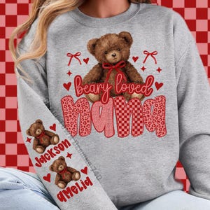 May include: A gray sweatshirt featuring a brown teddy bear with a red bow and the words "beary loved mama". The sleeve displays two teddy bears with the names "Jackson" and "Amelia".
