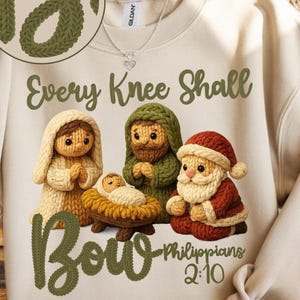 May include: Cream-colored sweatshirt featuring a knitted nativity scene with the text "Every Knee Shall Bow" and "Philippians 2:10." The design includes figures of Mary, Joseph, baby Jesus, and Santa Claus. The words "Faux Stitched" are at the top.
