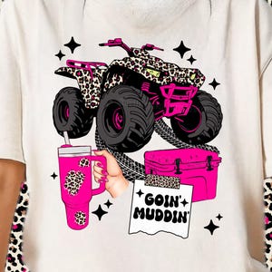 May include: Cream-coloured t-shirt featuring a pink ATV with leopard print accents, a pink tumbler, and a pink cooler. The text "GOIN' MUDDIN'" is on a torn paper. Black star graphics are scattered around the design.
