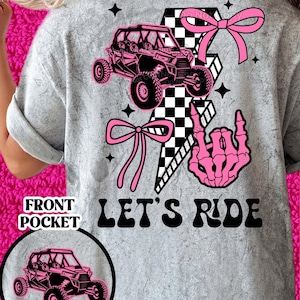 May include: Gray t-shirt with a pink and black checkered design, a pink bow, a skeleton hand, and a black and white image of an off-road vehicle. The text "Let's Ride" is printed in black on the shirt.