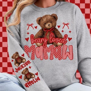 May include: A gray sweatshirt with a teddy bear graphic and the words "beary loved Mama". The design includes red hearts, bows, and the names "Jackson" and "Amelia" on the sleeve. The teddy bear has a red bow.