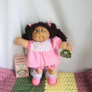 Puede incluir: Muñeca Cabbage Patch Kids con coletas marrones, vestida con un vestido rosa, bombachos y zapatos con ribete de encaje blanco. La muñeca tiene una etiqueta y un certificado de nacimiento. Sobre una colorida manta de ganchillo.