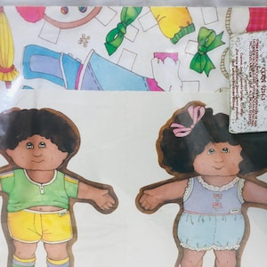 May include: Two Cabbage Patch Kids paper dolls. One doll wears a green shirt and yellow shorts, the other wears a purple top and blue shorts. The dolls have brown hair and are on a white background. The image includes text.