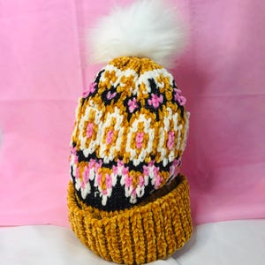 Peut inclure: Un bonnet en tricot jaune moutarde et noir avec un pompon en fausse fourrure blanche. Le bonnet pr&eacute;sente un motif g&eacute;om&eacute;trique blanc, rose et noir. Le bord est jaune moutarde uni, sur fond rose.