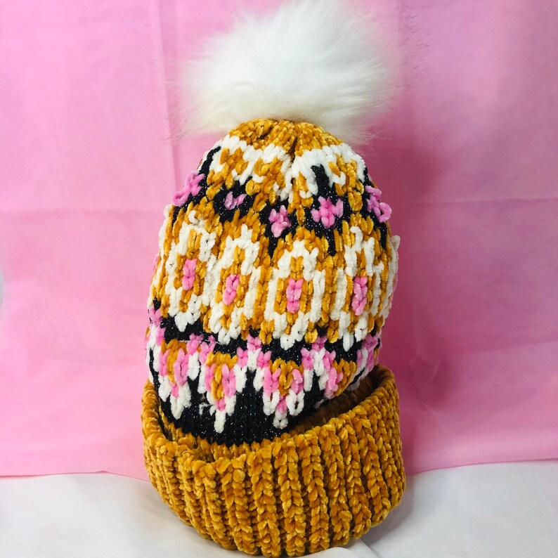 Peut inclure: Un bonnet en tricot jaune moutarde et noir avec un pompon en fausse fourrure blanche. Le bonnet pr&eacute;sente un motif g&eacute;om&eacute;trique blanc, rose et noir. Le bord est jaune moutarde uni, sur fond rose.