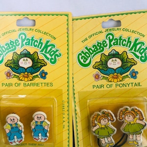 Könnte beinhalten: Zwei Vintage-Cabbage-Patch-Kids-Schmucksets auf gelber Verpackung. Ein Set ist ein Paar Haarspangen mit Jungenfiguren, das andere ein Paar Haargummis mit Mädchenfiguren. Die Verpackung enthält den Text "The Official Jewelry Collection."