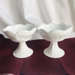 Puede incluir: Dos cuencos blancos de vidrio de leche con pedestal y diseño de uva. Los cuencos están sobre una superficie burdeos.