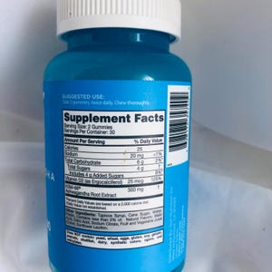 K&ouml;nnte beinhalten: Eine blaue Erg&auml;nzungsflasche mit wei&szlig;em Deckel. Das Etikett zeigt "N&auml;hrwertangaben" und Details zum Produkt, einschlie&szlig;lich Kalorien, Vitaminen und Ashwagandha-Wurzelextrakt. Die Flasche enth&auml;lt 30 Portionen.