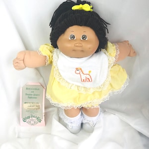 Puede incluir: Muñeca Cabbage Patch Kids con cabello oscuro y ojos marrones, vestida con un vestido amarillo con ribete de encaje y un babero blanco con un diseño de caballo. La muñeca tiene calcetines y zapatos blancos. Se incluye un certificado de nacimiento y papeles de adopción.